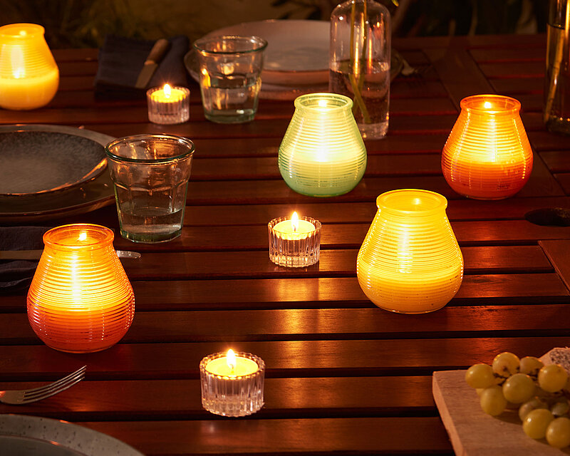 Sanfte Abendbeleuchtung auf der Terrasse: bunte Outdoor‑Kerzen und Citronella‑Kerzen in stilvollen Windlichtern erhellen den Holztisch und schaffen gemütliche, mückenfreie Stunden im Freien – ideal für Sommerabende mit Freunden.