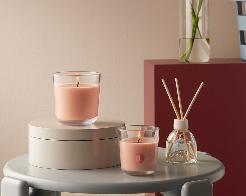 Das Bild zeigt eine stilvoll eingerichtete Szene mit zwei Duftkerzen im Glas sowie einem Raumduft-Diffuser aus der Bolsius True Scents Pfirsich-Serie. Die Kerzen verbreiten einen sanften, fruchtigen Pfirsichduft, der nicht nur für eine angenehme Atmosphäre sorgt, sondern auch gezielt unangenehme Tabakgerüche neutralisiert. Die harmonische Farbgebung in warmen Pfirsichtönen unterstreicht die entspannende Wirkung des Raumdufts. Ideal für Wohn- oder Schlafzimmer – für ein Zuhause, das frisch, einladend und rauchfrei duftet.