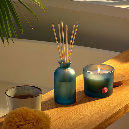 Ein entspannendes Badezimmer-Setting im sanften Tageslicht: Ein zart bläuliches Glasgefäß mit Duftstäbchen steht mittig auf einem rustikalen Holzboard über der schaumigen Badewanne. Daneben flackert eine Kerze im passenden Blauton. Das Arrangement umgibt Moos-Schwamm, Teebecher und seifige Accessoires, eingerahmt von Palmenblatt-Schatten und Jalousien-Licht – ganz auf die beruhigende Wirkung des Raumdufts von Bolsius ausgerichtet.