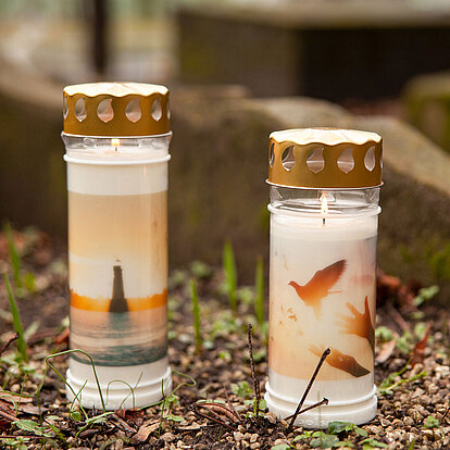 Zwei stilvolle Grabkerzen mit Deckel und Motiv stehen nebeneinander auf einem Grab. Die brennenden Gedenkkerzen zeigen emotionale Bildmotive – ein Leuchtturm bei Sonnenuntergang und fliegende Tauben mit ausgestreckten Händen. Der goldfarbene Deckel schützt die Flamme zuverlässig vor Wind, während die bedruckte Hülle eine würdevolle, persönliche Note verleiht. Ideal für eine langlebige und ausdrucksstarke Grabgestaltung.