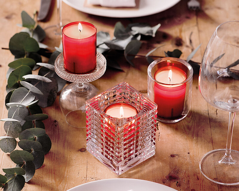 Auf einem rustikalen braunen Holztisch stehen zwei Teller mit Servietten, Messern und Gabeln sowie zwei Weingläser. Auf dem Tisch befinden sich mehrere Zweige Eukalyptus Cinerea und ein Bolsius Relight Halter Ribbed, einen Relight Halter Square und ein Relight Halter Moods mit je einem brennenden Nachfüller Relight mit rotem Cup.