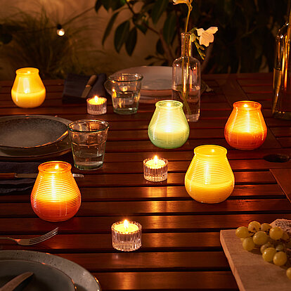 Sanfte Abendbeleuchtung auf der Terrasse: bunte Outdoor‑Kerzen und Citronella‑Kerzen in stilvollen Windlichtern erhellen den Holztisch und schaffen gemütliche, mückenfreie Stunden im Freien – ideal für Sommerabende mit Freunden.