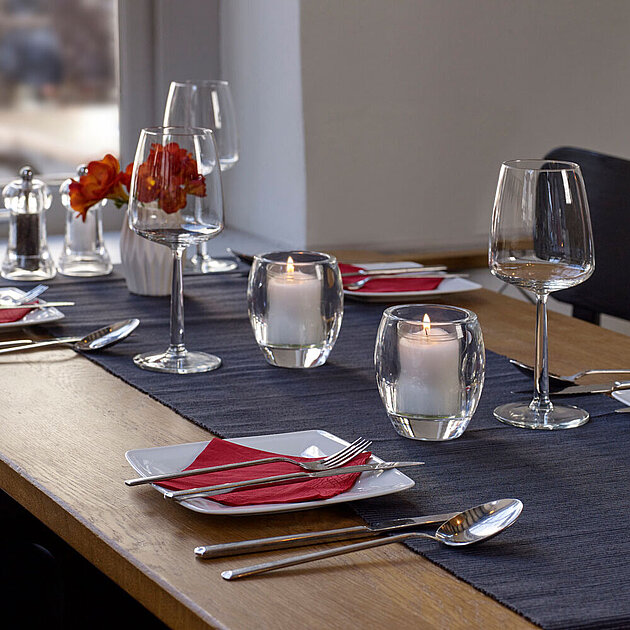 bolsius-kerzen-gastronomie-impression-2023-1 Die Kerzen Gastronomie setzt auf eine stilvolle Tischdekoration in einem Restaurant, die mit weißen Tellern und roten Servietten anspruchsvoll gestaltet ist. Auf dem Tisch steht eine weiße Vase mit roten Blumen und leere Weingläser, begleitet von zwei brennenden Relight Kerzen in einem ovalen Glashalter, die eine gemütliche und einladende Stimmung verbreiten.