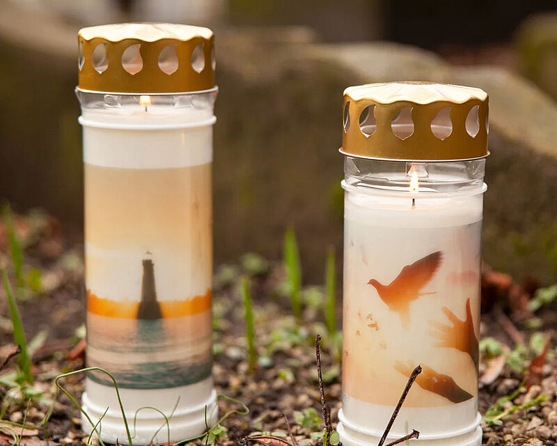 Zwei stilvolle Grabkerzen mit Deckel und Motiv stehen nebeneinander auf einem Grab. Die brennenden Gedenkkerzen zeigen emotionale Bildmotive – ein Leuchtturm bei Sonnenuntergang und fliegende Tauben mit ausgestreckten Händen. Der goldfarbene Deckel schützt die Flamme zuverlässig vor Wind, während die bedruckte Hülle eine würdevolle, persönliche Note verleiht. Ideal für eine langlebige und ausdrucksstarke Grabgestaltung.