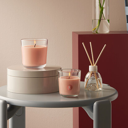 Das Bild zeigt eine stilvoll eingerichtete Szene mit zwei Duftkerzen im Glas sowie einem Raumduft-Diffuser aus der Bolsius True Scents Pfirsich-Serie. Die Kerzen verbreiten einen sanften, fruchtigen Pfirsichduft, der nicht nur für eine angenehme Atmosphäre sorgt, sondern auch gezielt unangenehme Tabakgerüche neutralisiert. Die harmonische Farbgebung in warmen Pfirsichtönen unterstreicht die entspannende Wirkung des Raumdufts. Ideal für Wohn- oder Schlafzimmer – für ein Zuhause, das frisch, einladend und rauchfrei duftet.