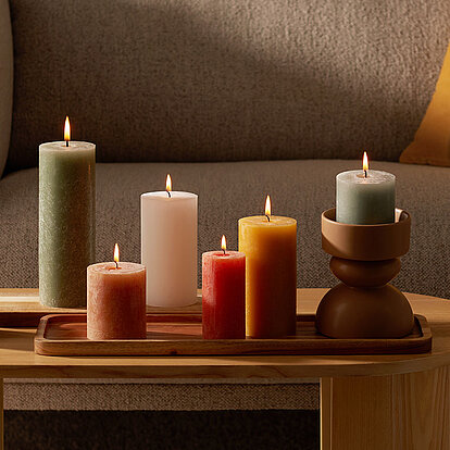 Bunte Stumpenkerzen in warmen Farben brennen stimmungsvoll auf einem Holztablett im Wohnzimmer. Die gemütliche Einrichtung mit Sofa, Kissen und Pflanzen sorgt für eine entspannte Atmosphäre. Die Kerzen in Grün, Rot, Orange, Weiß und Grau sind farblich stimmungsvoll aufeinander abgestimmt.