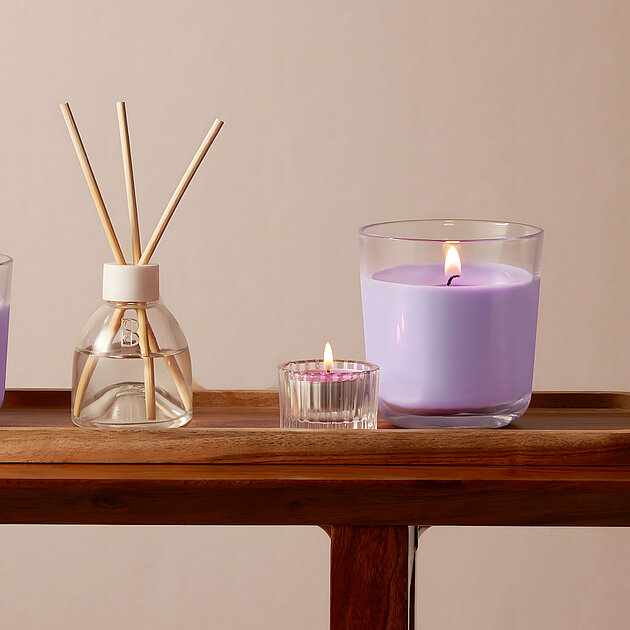 Auf einem schmalen Holzregal stehen mehrere Bolsius Duft‑Teelichter in zartem Lavendelfarbton – einige glimmen bereits und verbreiten sanfte Lichtakzente. Dazwischen befindet sich ein diffuser Raumduftspender mit Stäbchen, der das Duftbild vervollständigt. Der florale Lavendelduft wird ergänzt von harmonischer Dekoration: ein rosa Wellen-Vase mit schlanker Einzelblüte, darunter eine kleine Buch‑Miniatur und eine stilvolle Hocker-Oberfläche in Blau. Diese stilvolle Inszenierung lenkt voll auf die Duft-Teelichter und schafft ein ruhiges, aromatisches Ambiente.