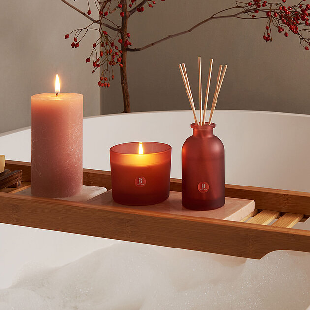 Eine sanft brennende Bolsius Duftkerze im Glas strahlt warmes Licht über eine freistehende Badewanne aus, begleitet von einem passenden Raumduft-Diffuser in stilvollem Rot. Eine rustikale Stumpenkerze ergänzt das Arrangement auf einem Holz-Wannenbrett, während nebenbei Wasser leise aus dem Armatur perlt. Im Hintergrund sorgen zarte Zweige mit roten Beeren für einen dezenten, natürlichen Touch – ideal für entspannende Wellnessmomente mit dem intensiven Aroma der Bolsius Duftkerzen.